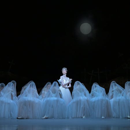 A ballet dancer from the National Ballet of Japan stands in the centre of the stage with her arms slightly crossed in front of her waist. She wears a white dress, small translucent wings and a crystal tiara on her head. She is encircled by dozens of dancers kneeling at her feet. They wear white dresses, small wings and white translucent veils that drape over their heads giving them a ghostly appearance. The stage set is minimal with a full moon in the night sky and soft blue lighting.