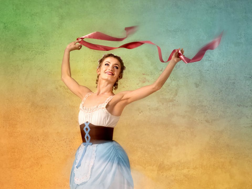 Anna Rose O'Sullivan dressed in the costume for Lise in La Fille mal gardée. She wears a costume with a white bodice, light blue apron skirt, and brown corset and dances en pointe against a softly textured, multicolored backdrop. She holds a pink ribbons aloft, forming graceful arcs in the air as she smiles and strikes a joyful pose.