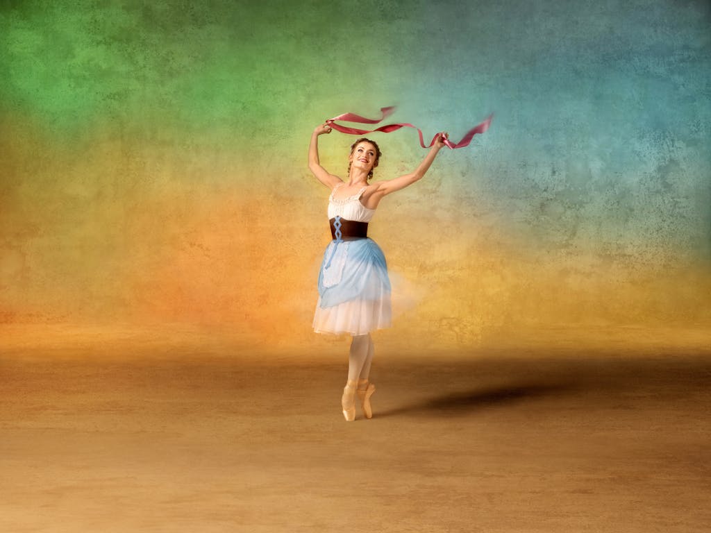 Anna Rose O'Sullivan dressed in the costume for Lise in La Fille mal gardée. She wears a costume with a white bodice, light blue apron skirt, and brown corset and dances en pointe against a softly textured, multicolored backdrop. She holds a pink ribbons aloft, forming graceful arcs in the air as she smiles and strikes a joyful pose.