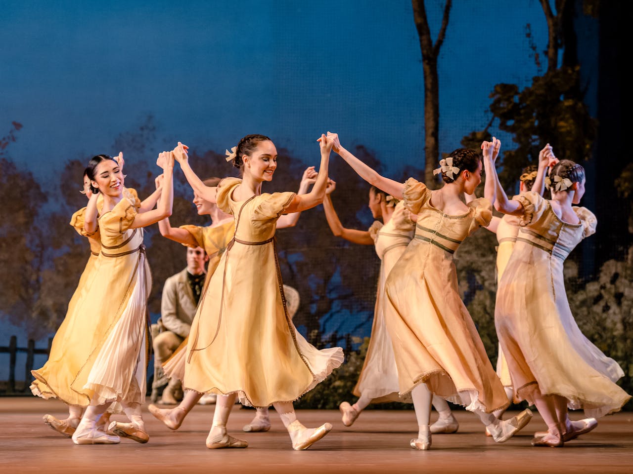 A group of dancers of The Royal Ballet in flowing yellow dresses performs a Onegin on stage, holding hands in a circular formation. They wear ballet slippers and have ribbons in their hair. The background features a painted outdoor scene with trees and a wooden fence, creating a romantic atmosphere.
