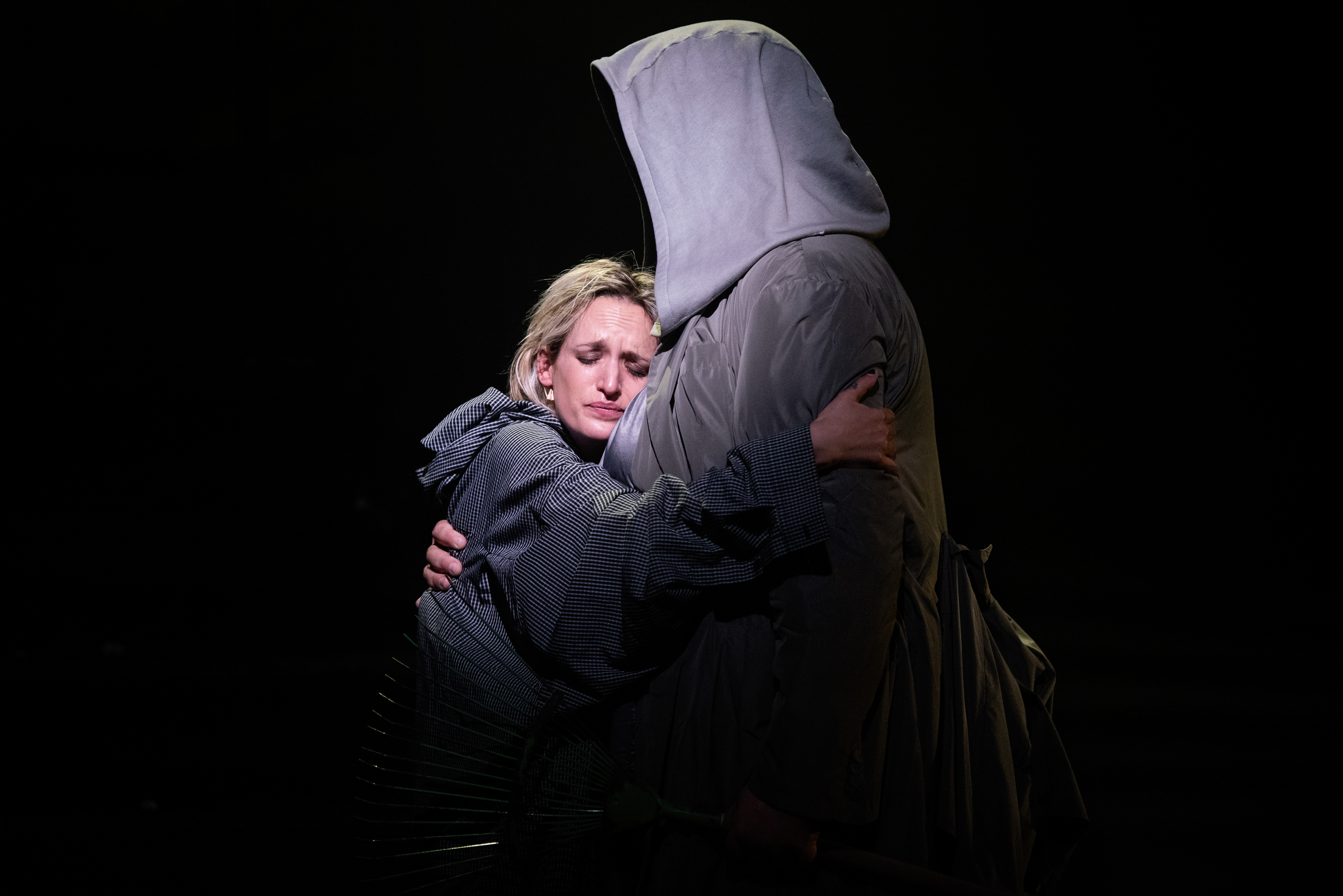 Agathe Rousselle performs as Blake in Oliver Leith’s production of Last Days. She shares a hug with a performer who is wearing a large hood that covers their face. She leans into the embrace with an upset yet relieved expression.