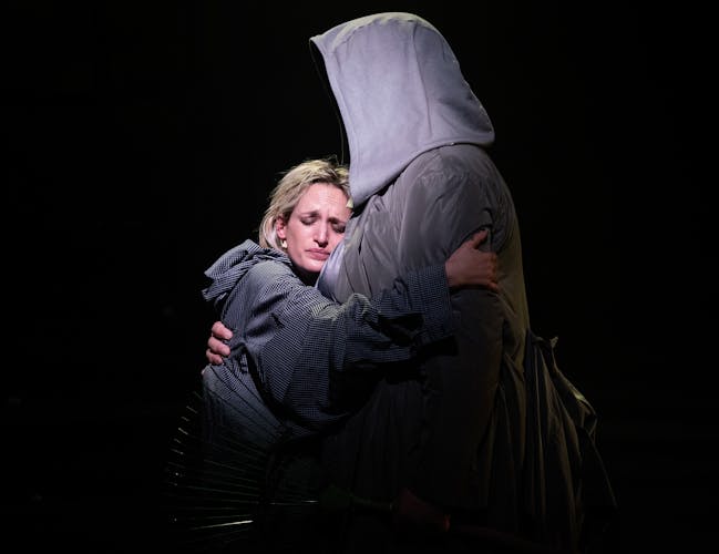 Agathe Rousselle performs as Blake in Oliver Leith’s production of Last Days. She shares a hug with a performer who is wearing a large hood that covers their face. She leans into the embrace with an upset yet relieved expression.