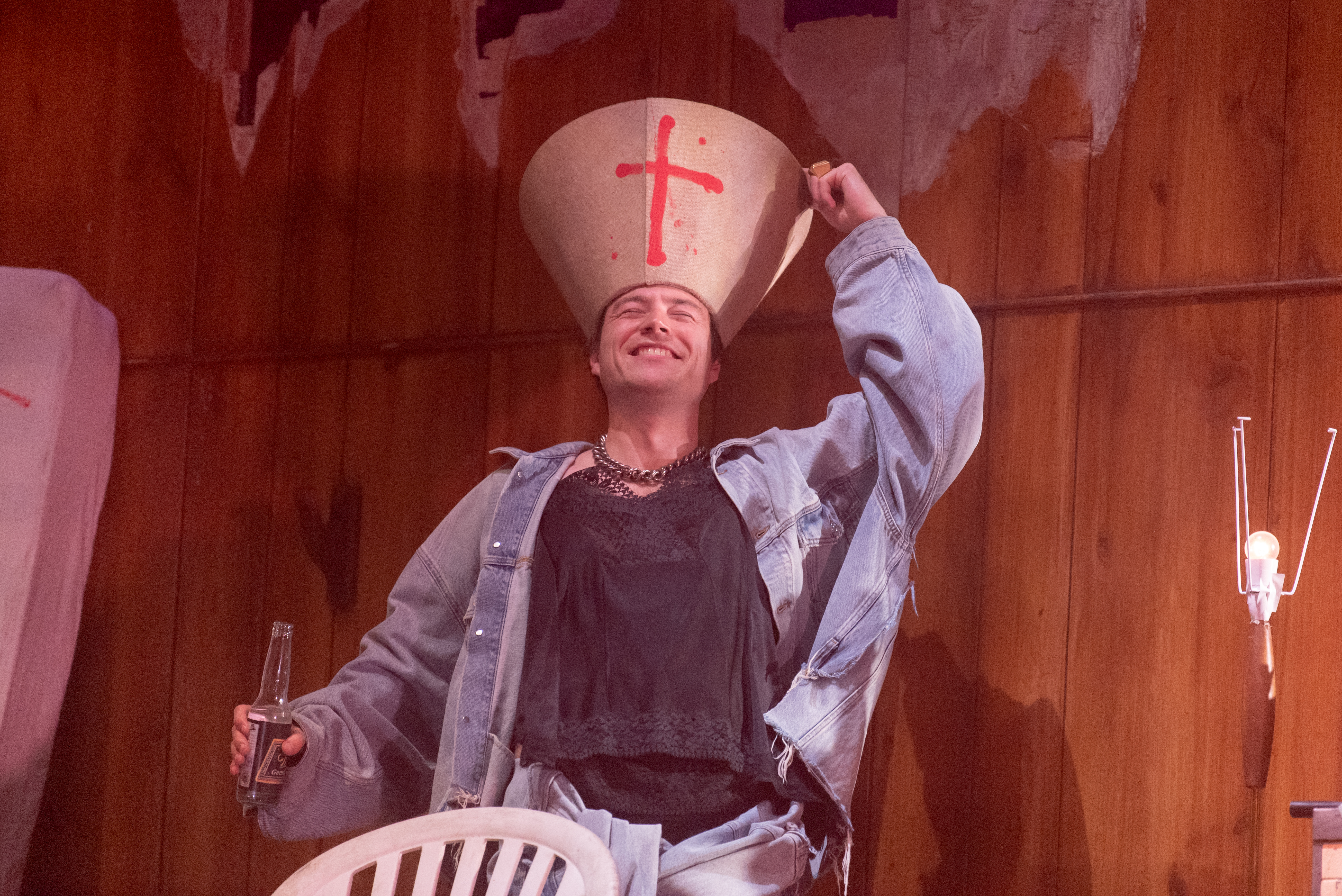 Seumas Begg performs as a housemate in Oliver Leith's Last Days. He is wearing an oversized denim jacket and a loose black t-shirt. In one hand he holds a bottle of beer, with the other he places an overturned lampshade on his head. The beige lampshade has a red cross spray painted on it. He tilts his head backwards and grins with his eyes closed, appearing to be intoxicated. 