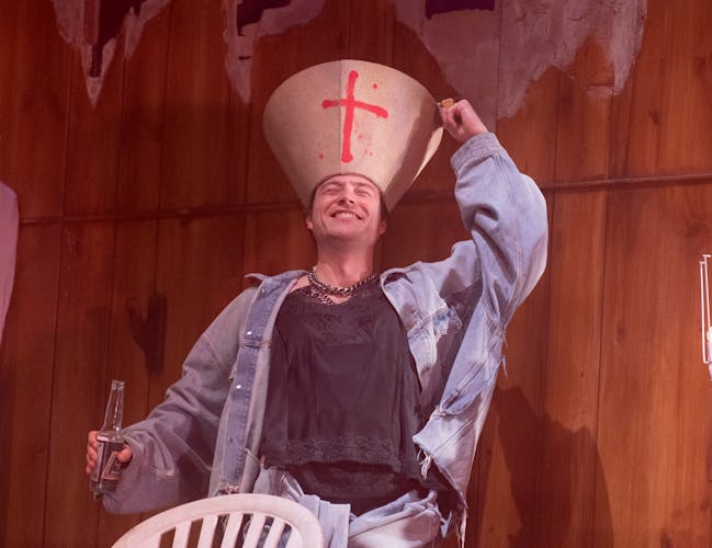 Seumas Begg performs as a housemate in Oliver Leith's Last Days. He is wearing an oversized denim jacket and a loose black t-shirt. In one hand he holds a bottle of beer, with the other he places an overturned lampshade on his head. The beige lampshade has a red cross spray painted on it. He tilts his head backwards and grins with his eyes closed, appearing to be intoxicated.