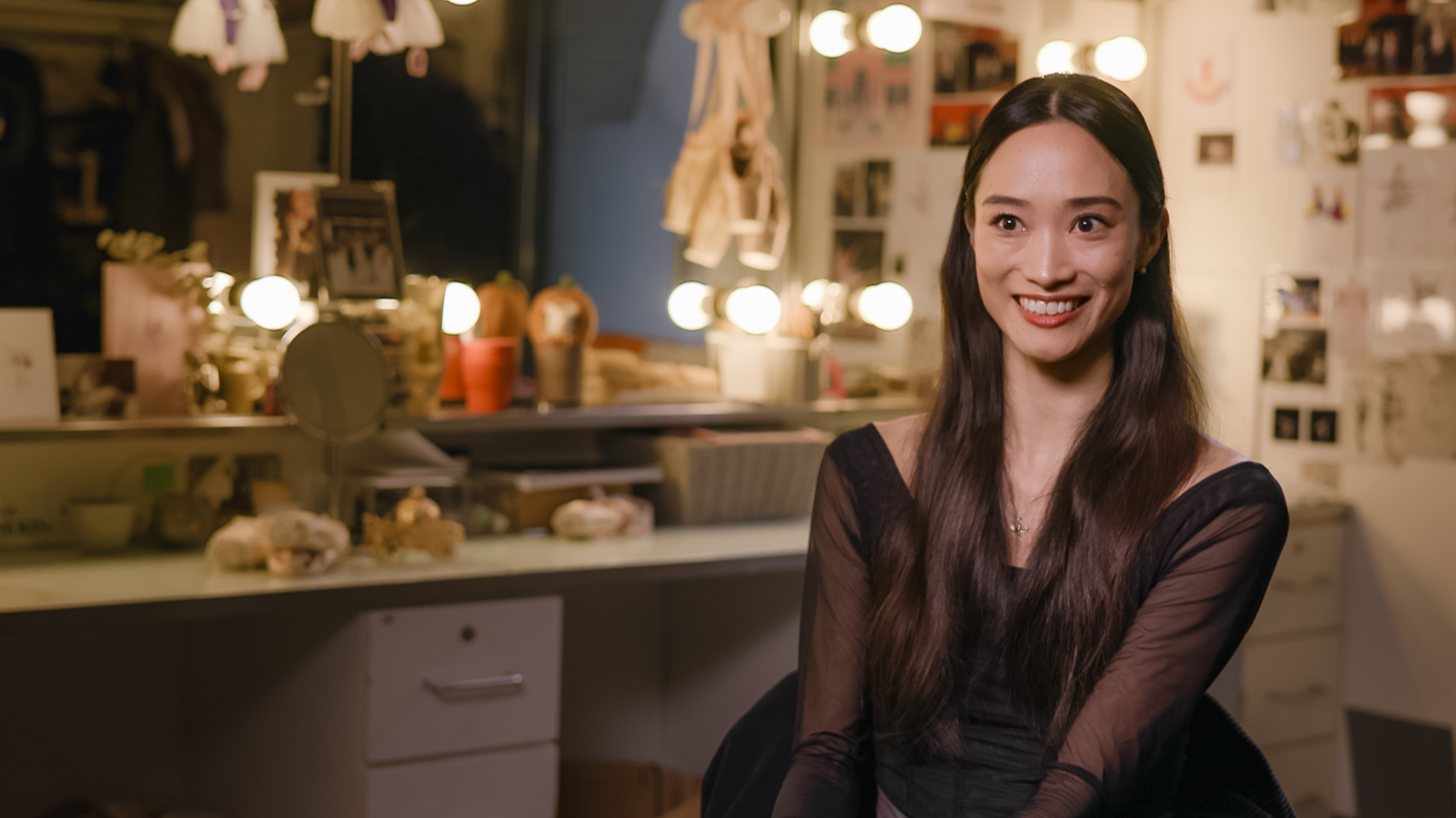 Principal Dancer Fumi Kaneko sits in a dressing room. In the background there is a mirror surrounded by lights. A pair of pale pink ballet shoes hang from the mirror.