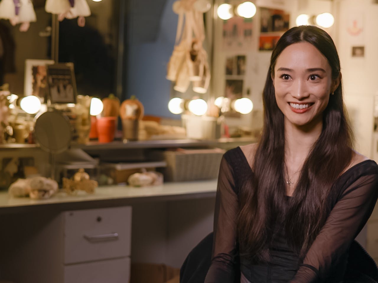 Principal Dancer Fumi Kaneko sits in a dressing room. In the background there is a mirror surrounded by lights. A pair of pale pink ballet shoes hang from the mirror.