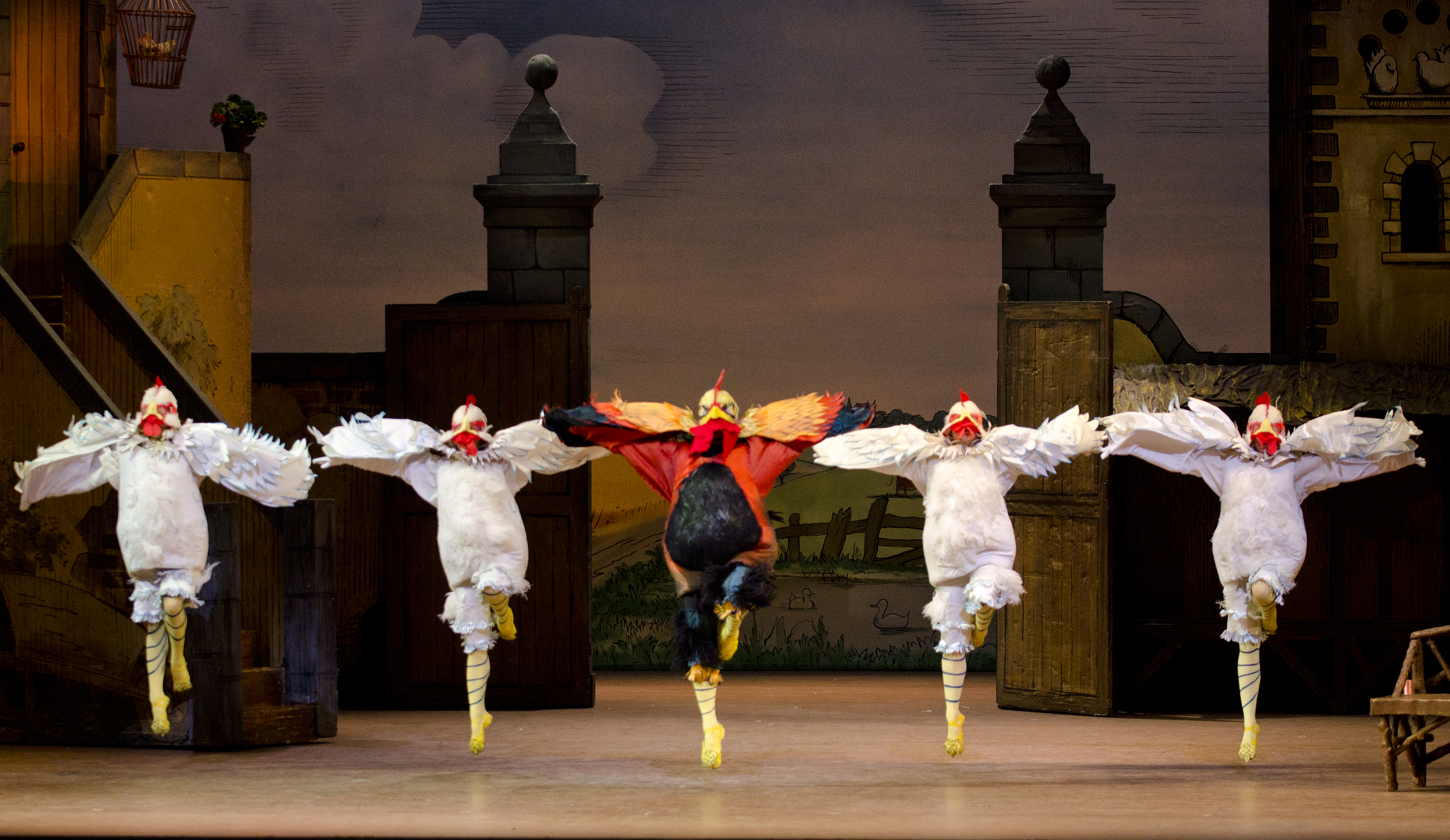 Five ballet dancers dressed as birds form a line across the stage. The central dancer has red, orange and black feathers. The two dancers on either side wear white feathers. All dancers hold the same position, leaping into the air with their left leg bent. Their arms look like wings and are outstretched. 