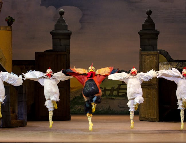 Five ballet dancers dressed as birds form a line across the stage. The central dancer has red, orange and black feathers. The two dancers on either side wear white feathers. All dancers hold the same position, leaping into the air with their left leg bent. Their arms look like wings and are outstretched.
