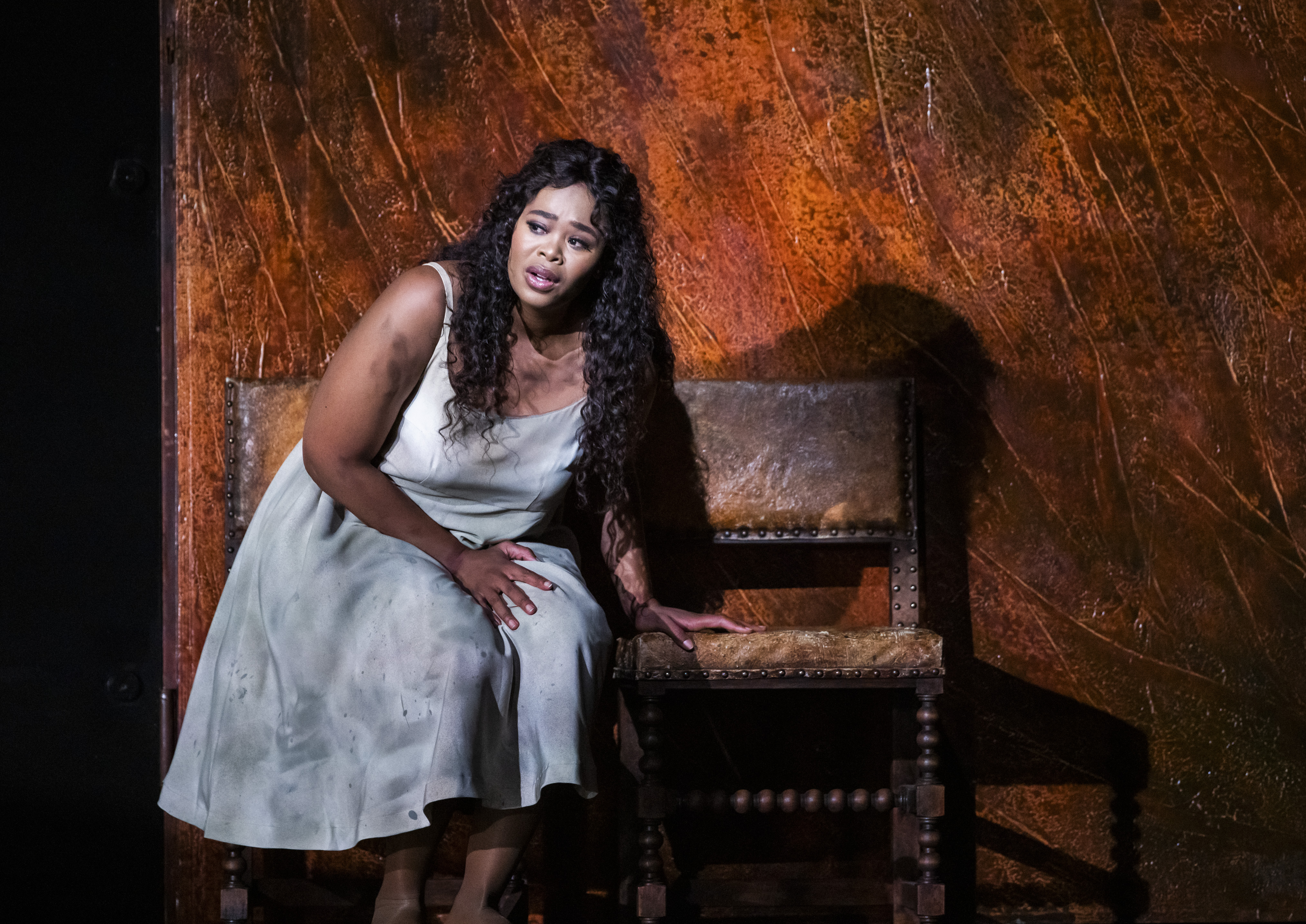 Opera singer Pretty Yende performs as Gilda in the Royal Opera’s production of Rigoletto. She sits on an old brown leather chair and leans on the chair next to her. She wears a plain white dress that appears dirty and stained.