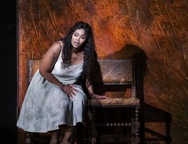 Opera singer Pretty Yende performs as Gilda in the Royal Opera’s production of Rigoletto. She sits on an old brown leather chair and leans on the chair next to her. She wears a plain white dress that appears dirty and stained.