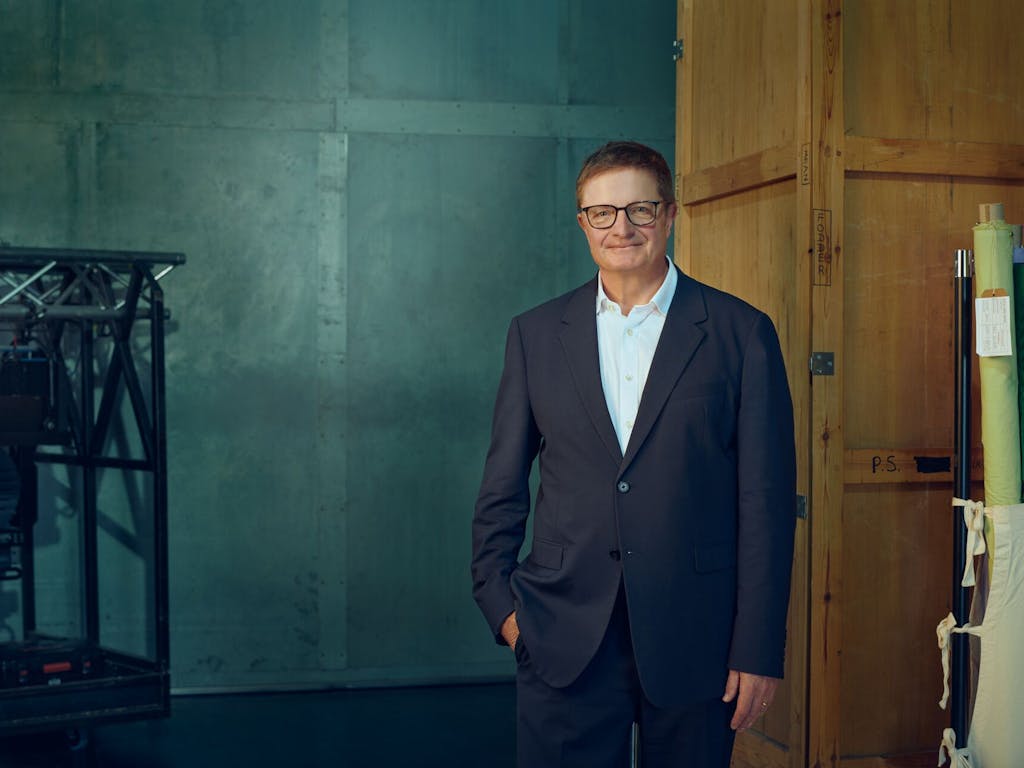 The Royal Ballet and Opera CEO Alex Beard stands in a room with lighting rigs, big wooden doors, and rolls of fabric in a bag. He wears a dark blue suit, light blue shirt and black framed glasses.