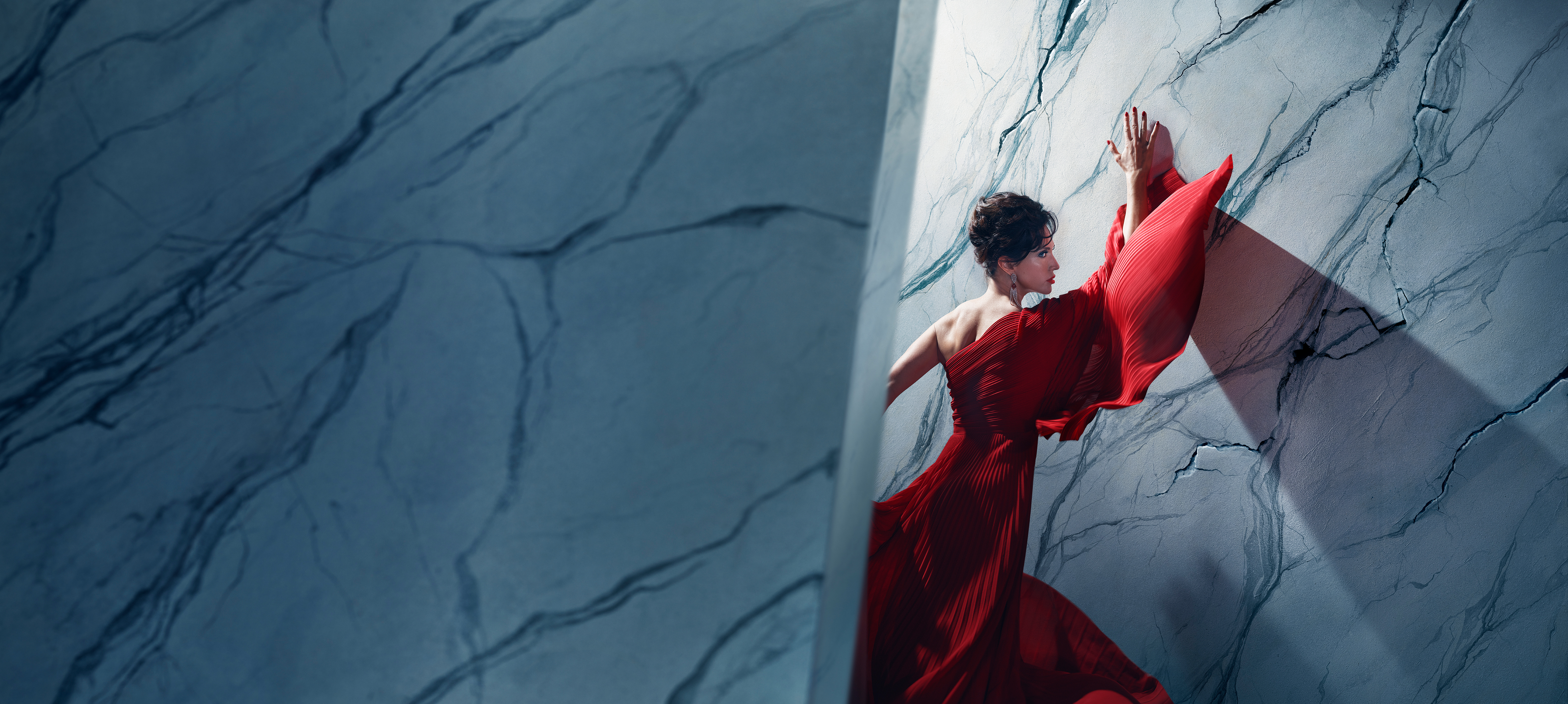 An actor playing Tosca in Oliver Mears’ production of Tosca stands in a dramatic red gown against a marble-textured wall. Her hands are pressed above her head and her body angled towards it, while parts of the wall appear to be cracking around her. A strong diagonal shadow casts across the wall, adding depth and tension to the composition. The setting is abstract and theatrical, with cool-toned lighting contrasting against the flowing red fabric.