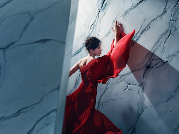An actor playing Tosca in Oliver Mears’ production of Tosca stands in a dramatic red gown against a marble-textured wall. Her hands are pressed above her head and her body angled towards it, while parts of the wall appear to be cracking around her. A strong diagonal shadow casts across the wall, adding depth and tension to the composition. The setting is abstract and theatrical, with cool-toned lighting contrasting against the flowing red fabric.