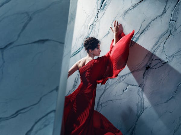 An actor playing Tosca in Oliver Mears’ production of Tosca stands in a dramatic red gown against a marble-textured wall. Her hands are pressed above her head and her body angled towards it, while parts of the wall appear to be cracking around her. A strong diagonal shadow casts across the wall, adding depth and tension to the composition. The setting is abstract and theatrical, with cool-toned lighting contrasting against the flowing red fabric.