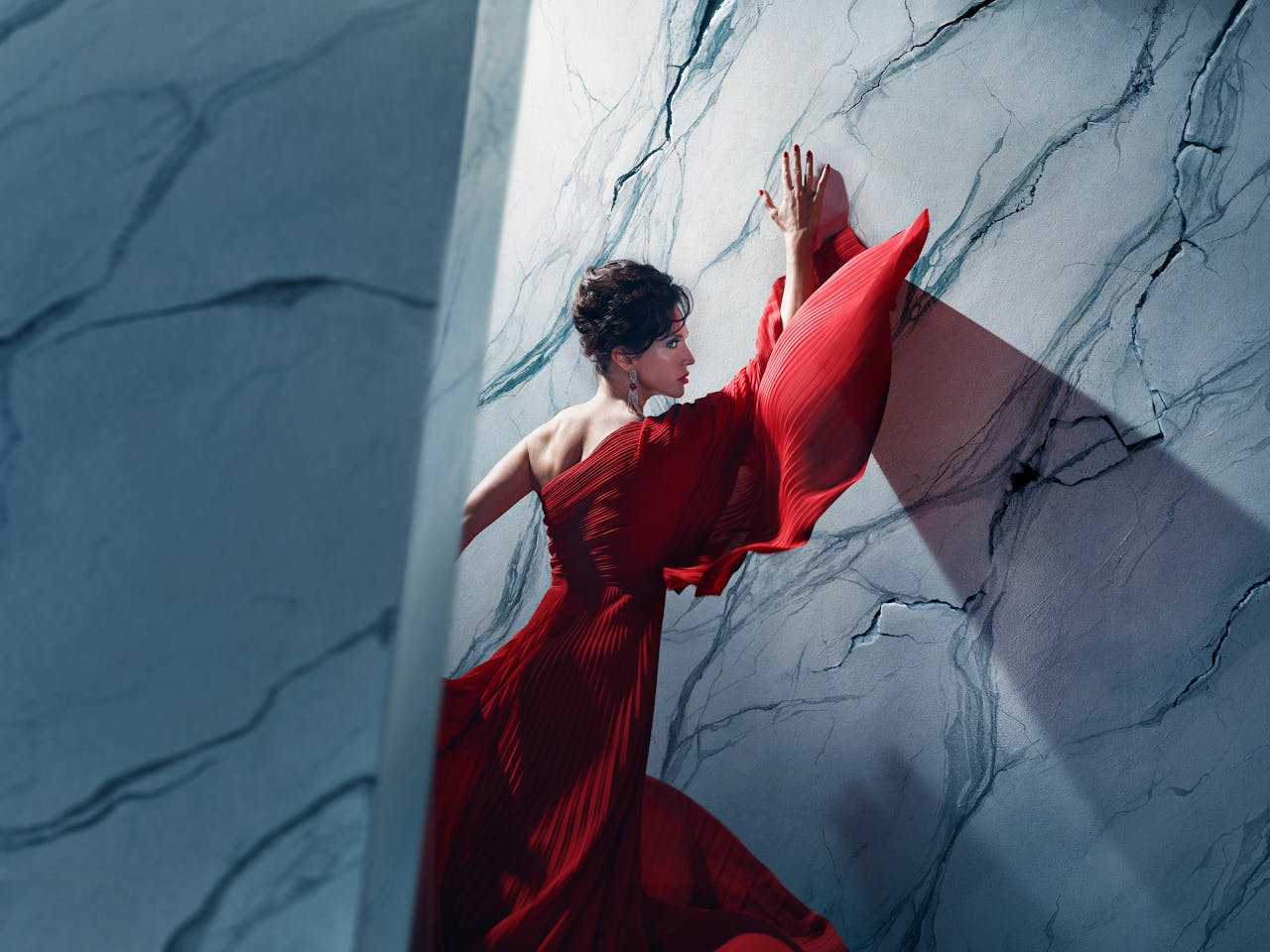An actor playing Tosca in Oliver Mears’ production of Tosca stands in a dramatic red gown against a marble-textured wall. Her hands are pressed above her head and her body angled towards it, while parts of the wall appear to be cracking around her. A strong diagonal shadow casts across the wall, adding depth and tension to the composition. The setting is abstract and theatrical, with cool-toned lighting contrasting against the flowing red fabric.