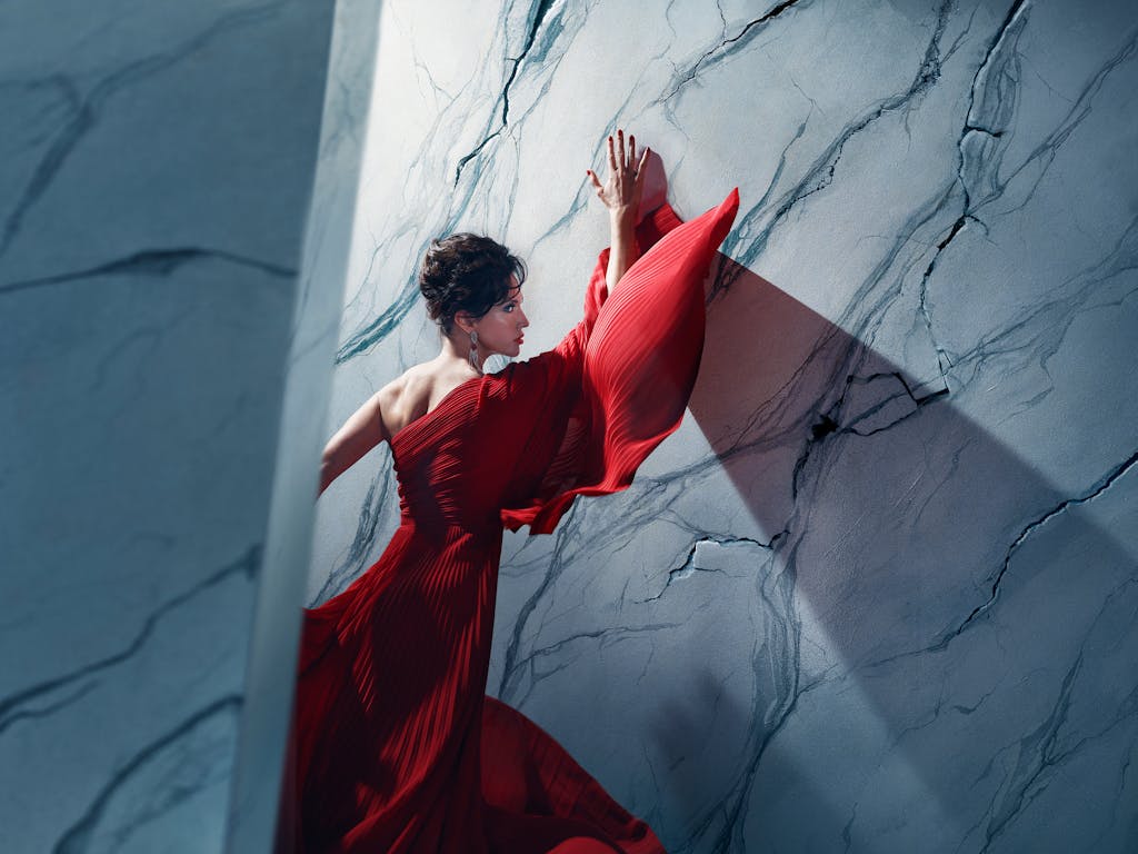 An actor playing Tosca in Oliver Mears’ production of Tosca stands in a dramatic red gown against a marble-textured wall. Her hands are pressed above her head and her body angled towards it, while parts of the wall appear to be cracking around her. A strong diagonal shadow casts across the wall, adding depth and tension to the composition. The setting is abstract and theatrical, with cool-toned lighting contrasting against the flowing red fabric.