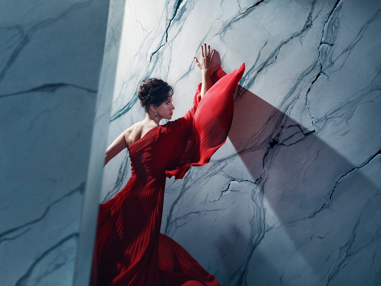 An actor playing Tosca in Oliver Mears’ production of Tosca stands in a dramatic red gown against a marble-textured wall. Her hands are pressed above her head and her body angled towards it, while parts of the wall appear to be cracking around her. A strong diagonal shadow casts across the wall, adding depth and tension to the composition. The setting is abstract and theatrical, with cool-toned lighting contrasting against the flowing red fabric.