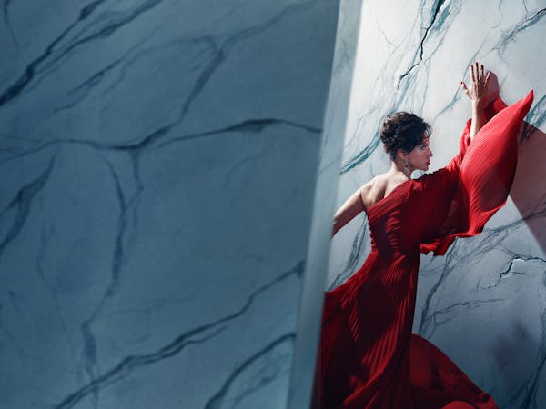 An actor playing Tosca in Oliver Mears’ production of Tosca stands in a dramatic red gown against a marble-textured wall. Her hands are pressed above her head and her body angled towards it, while parts of the wall appear to be cracking around her. A strong diagonal shadow casts across the wall, adding depth and tension to the composition. The setting is abstract and theatrical, with cool-toned lighting contrasting against the flowing red fabric.