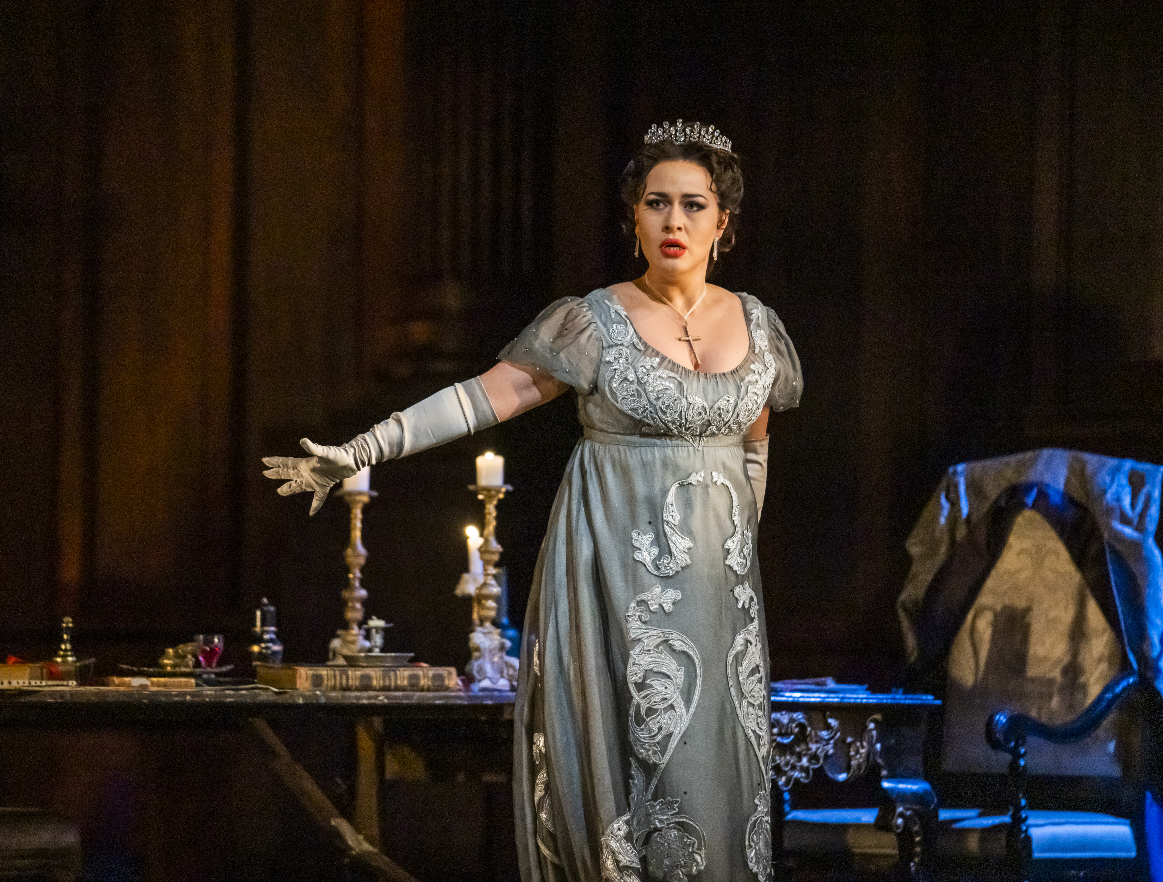 Soprano Elena Stikhina performs as Floria Tosca in Tosca. She stands in front of a lavish dinner table with her right arm outstretched. She is wearing a blue, embellished empire line dress, a jewelled tiara, elbow length gloves and a cross necklace. 