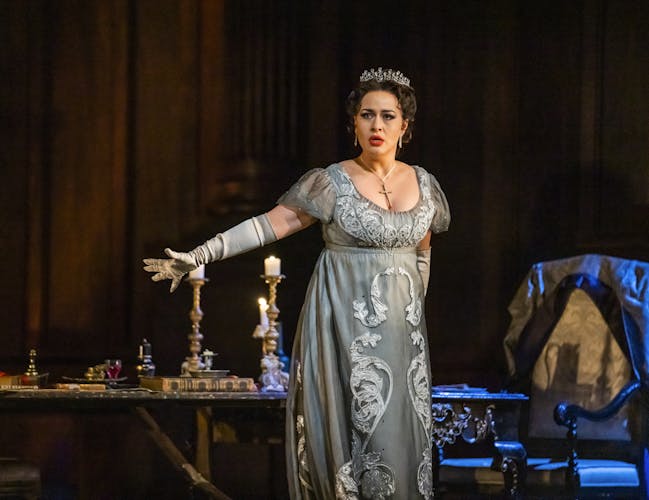 Soprano Elena Stikhina performs as Floria Tosca in Tosca. She stands in front of a lavish dinner table with her right arm outstretched. She is wearing a blue, embellished empire line dress, a jewelled tiara, elbow length gloves and a cross necklace.