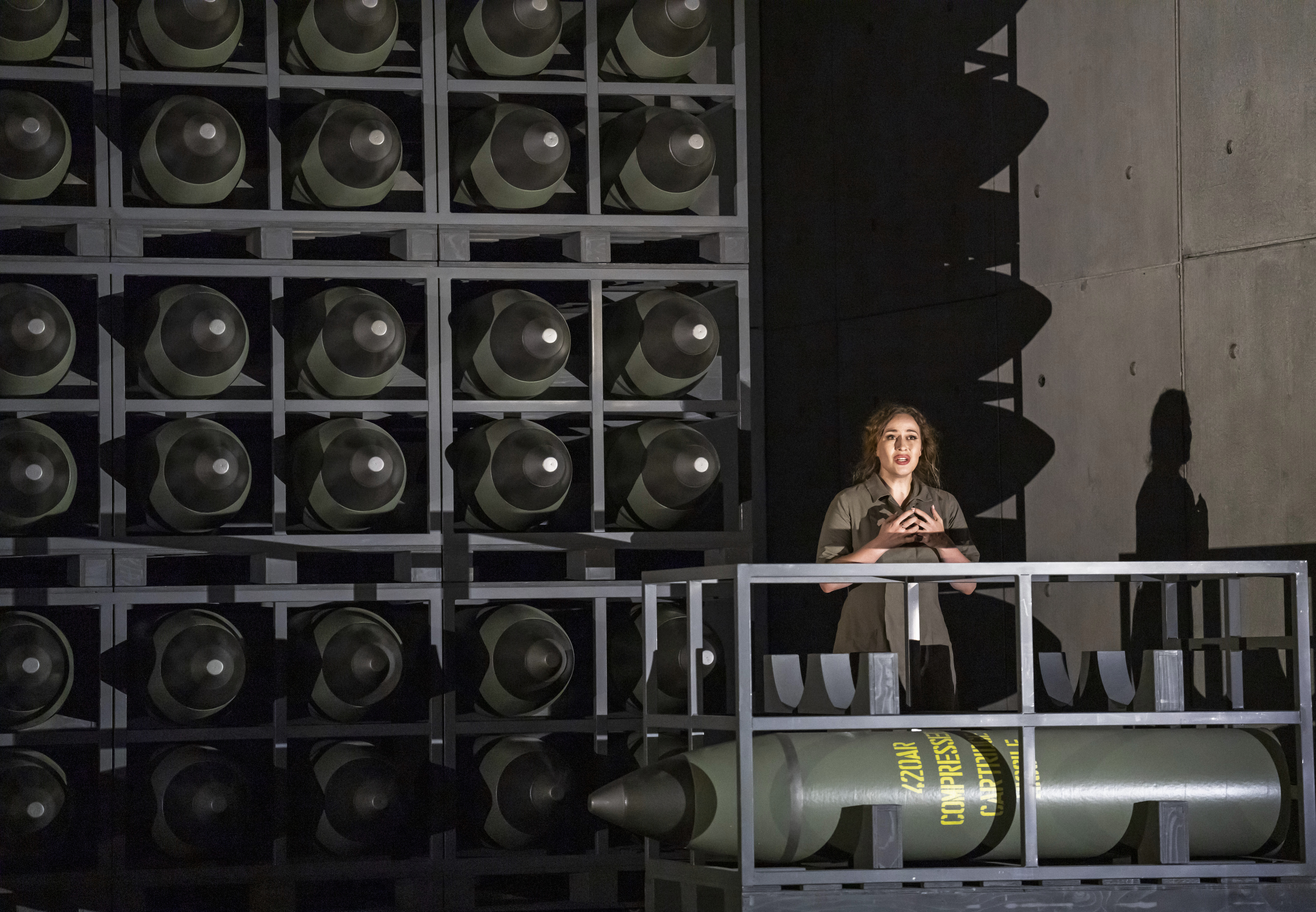 Soprano Elena Stikhina performs in Aida. She is wearing khaki military clothing and clasps her hands to her chest as she looks out to the audience. The stage resembles a grey, concrete bunker lined with large missiles in storage racks. 