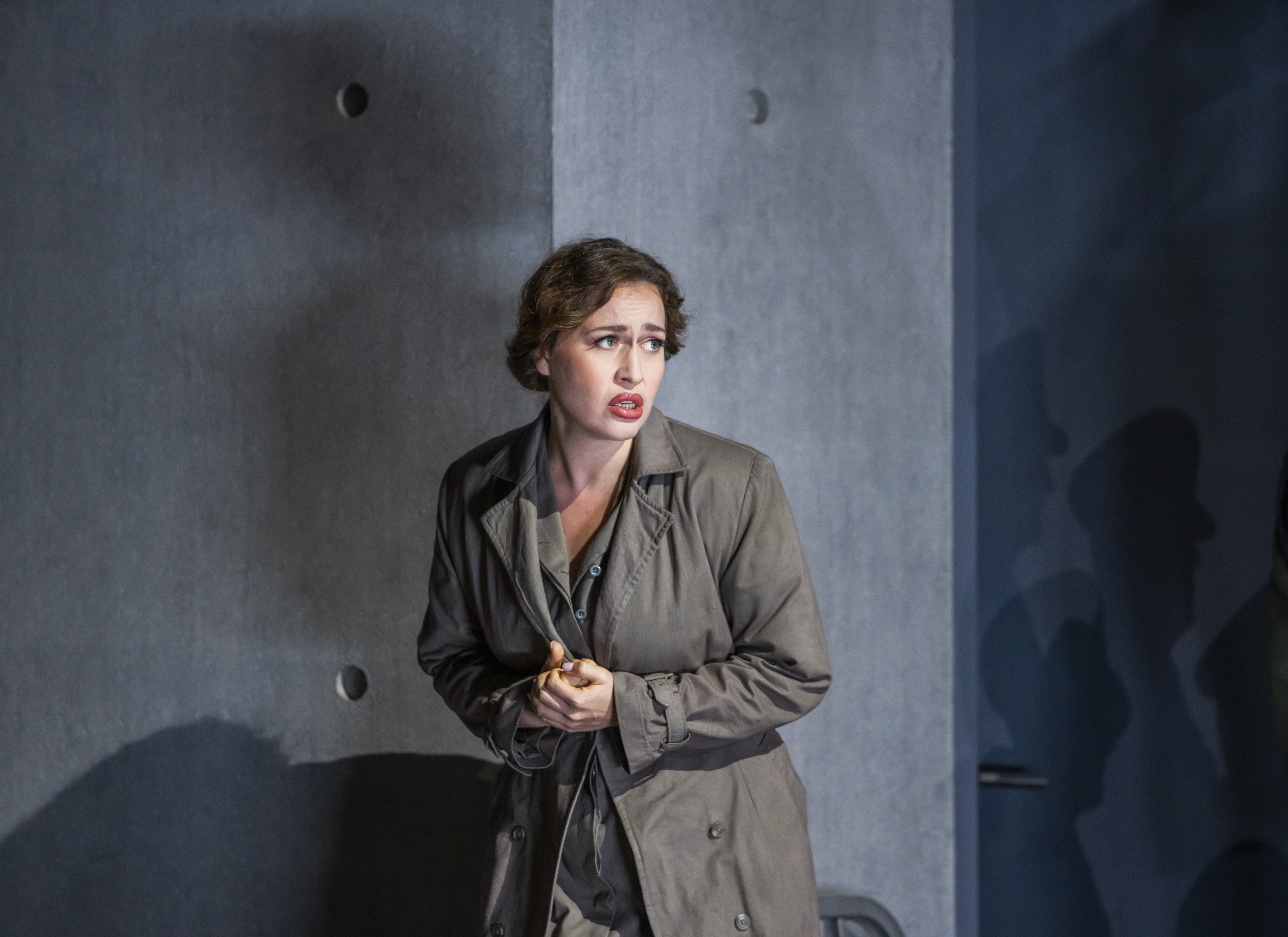 Soprano Elena Stikhina performs in Aida. She is dressed in grey military clothing with her hair tied up. She clasps her hands to her chest and gazes to the right with an anguished expression. The stage resembles a grey concrete bunker and shadows of figures, seemingly wearing military hats, loom on the walls behind her. 