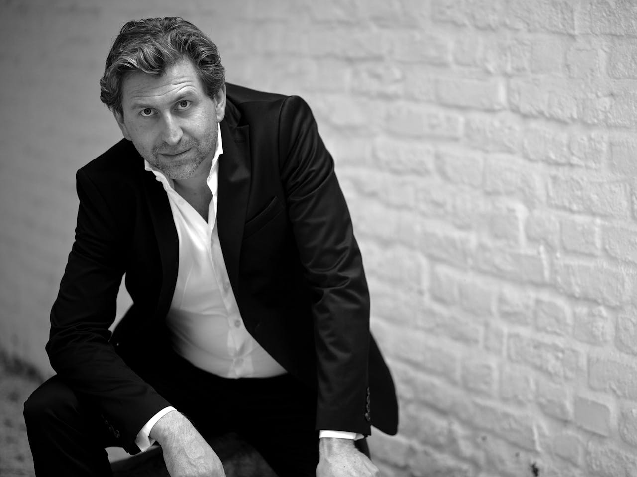 A black and white headshot of tenor Andreas Schager, who wears a black suit with a white shirt. He sits against an exposed brick wall that is painted white.
