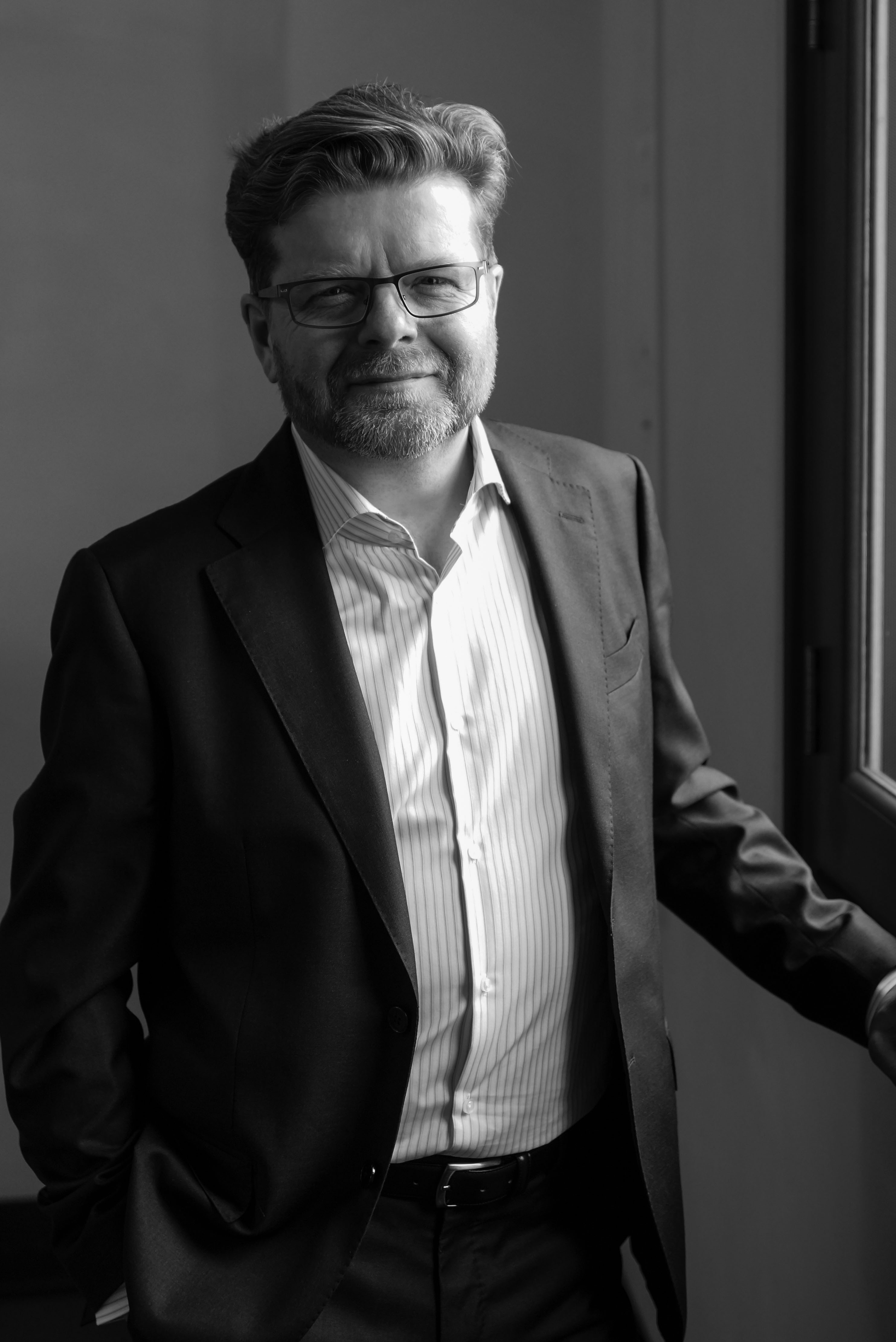 A black and white headshot of the opera singer Peter Hoare. He stands in a well-lit room and wears a black suit with white shirt.