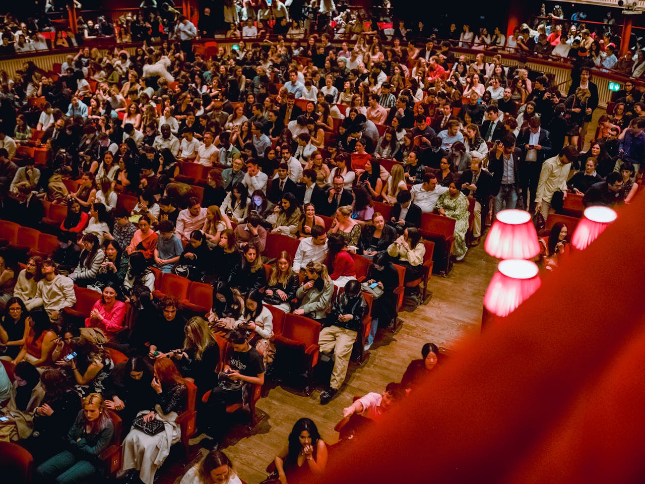 Many people are moving around the orchestra stalls area of a busy theatre. Some are sitting on red velvet theatre seats while others are moving in the space. It is the Main Stage of The Royal Opera House.