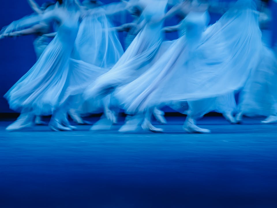 Blurred motion photograph of dancers of The Royal Ballet in flowing white dresses performing Balanchine’s Serenade on a dimly lit stage. The dancers' movements create a dreamy and ethereal effect, with their skirts billowing as they glide across the floor. The soft blue lighting enhances the sense of grace and fluidity in the performance.