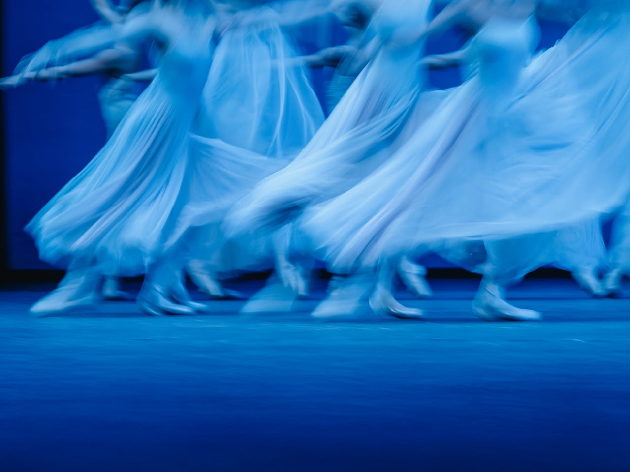 Blurred motion photograph of dancers of The Royal Ballet in flowing white dresses performing Balanchine’s Serenade on a dimly lit stage. The dancers' movements create a dreamy and ethereal effect, with their skirts billowing as they glide across the floor. The soft blue lighting enhances the sense of grace and fluidity in the performance.