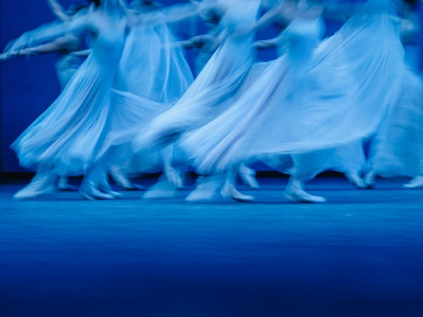 Blurred motion photograph of dancers of The Royal Ballet in flowing white dresses performing Balanchine’s Serenade on a dimly lit stage. The dancers' movements create a dreamy and ethereal effect, with their skirts billowing as they glide across the floor. The soft blue lighting enhances the sense of grace and fluidity in the performance.
