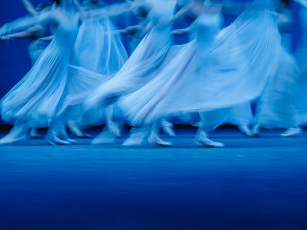 Blurred motion photograph of dancers of The Royal Ballet in flowing white dresses performing Balanchine’s Serenade on a dimly lit stage. The dancers' movements create a dreamy and ethereal effect, with their skirts billowing as they glide across the floor. The soft blue lighting enhances the sense of grace and fluidity in the performance.