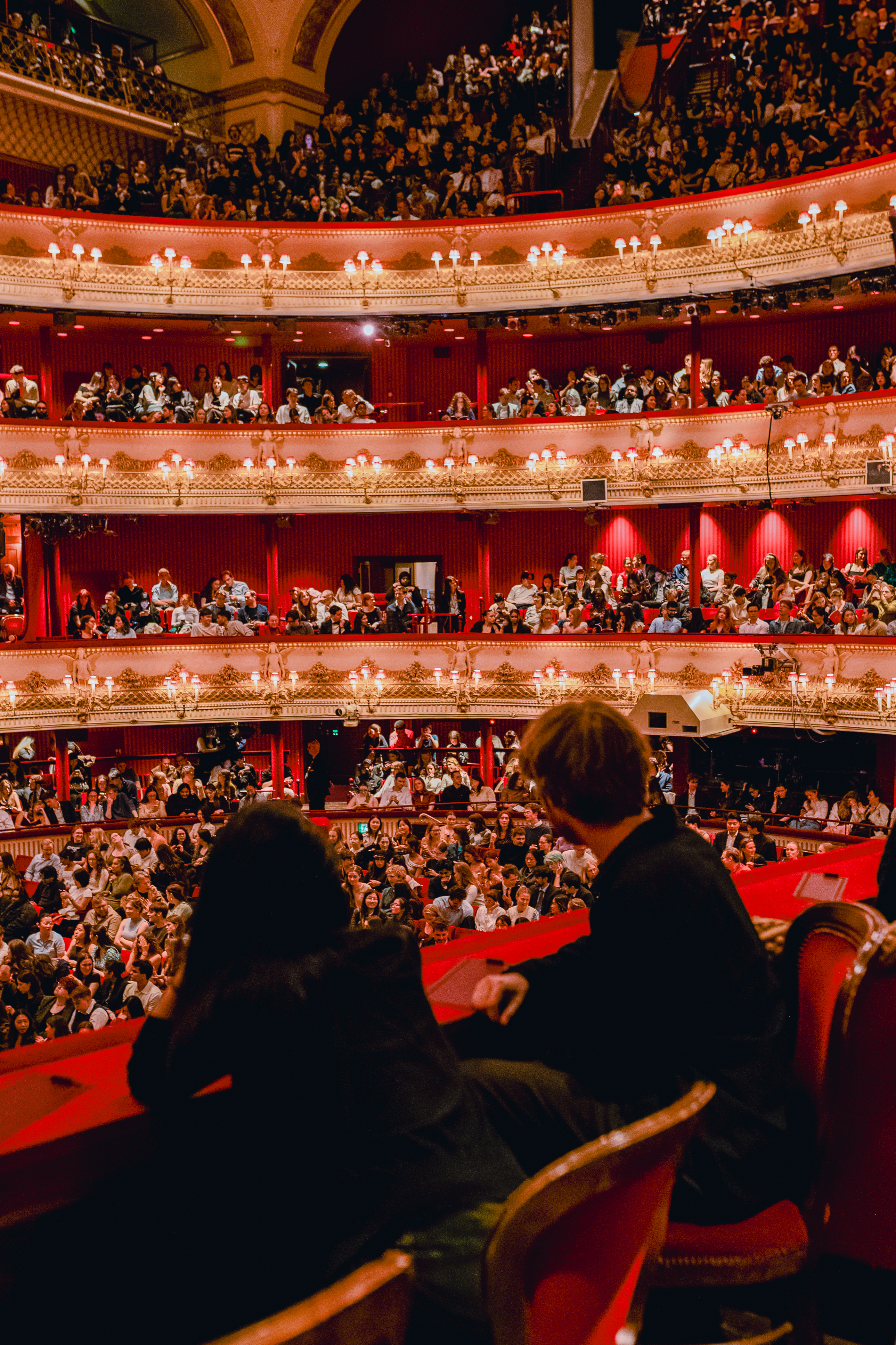 Two people wearing black look out from a balcony seat on the brightly lit full Main Stage auditorium of the Royal Opera House.