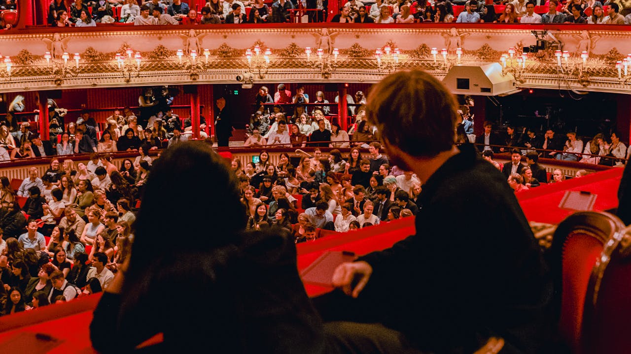 Two people wearing black look out from a balcony seat on the brightly lit full Main Stage auditorium of the Royal Opera House.