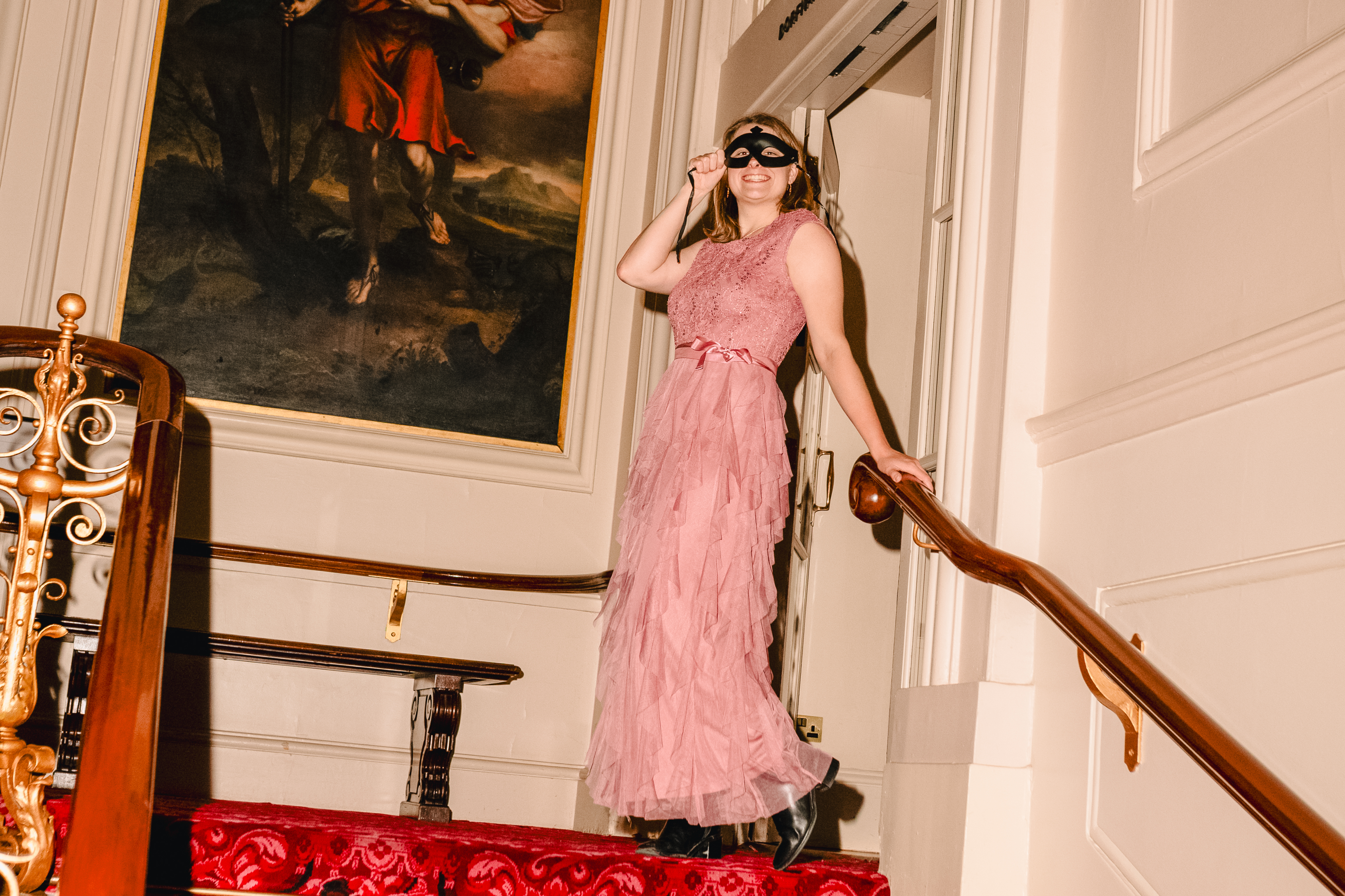 A person in a pink dress and black ornate eye mask stands at the top of red carpeted stairs holding the bannister. Behind them in a large traditional style portrait oil painting.