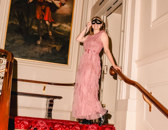 A person in a pink dress and black ornate eye mask stands at the top of red carpeted stairs holding the bannister. Behind them in a large traditional style portrait oil painting.