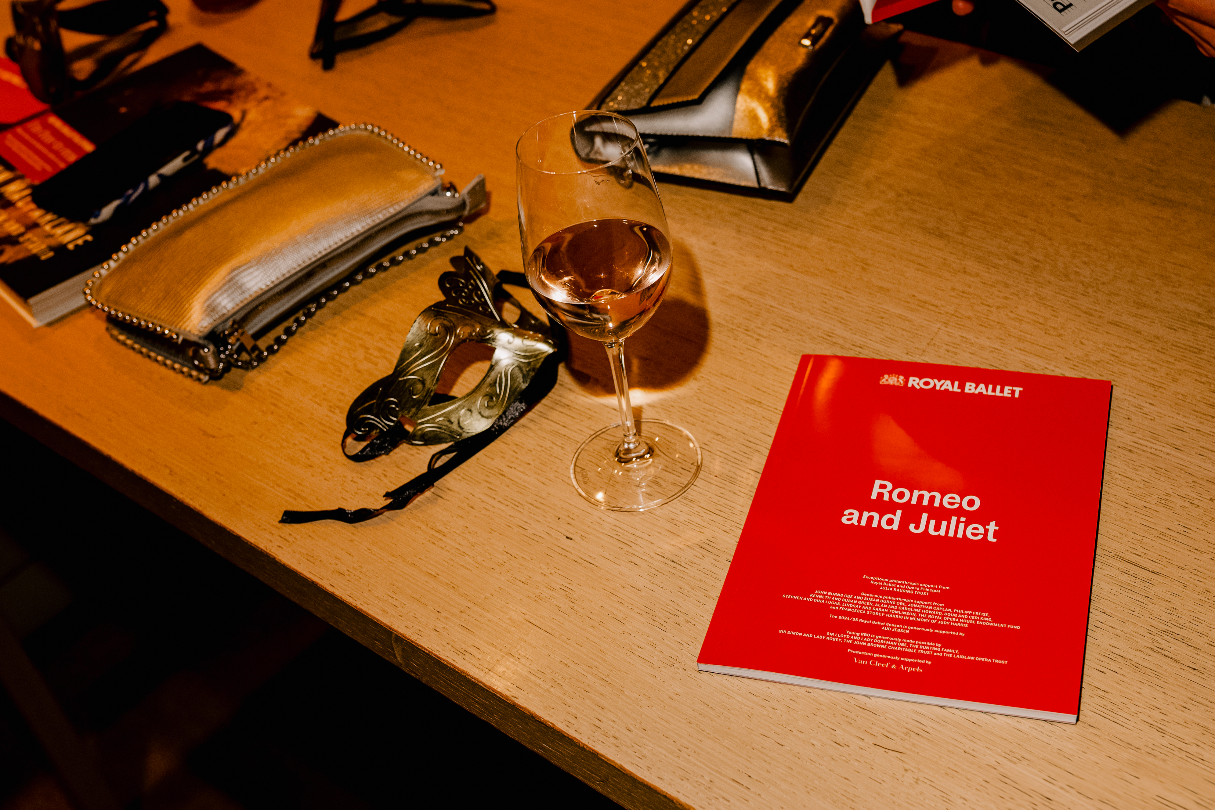 A red ballet programme for The Royal Ballet's production of Romeo and Juliet sits on a wooden table next to a wine glass of pink liquid, an ornate gold ribboned eye mask and two gold clutch bags.