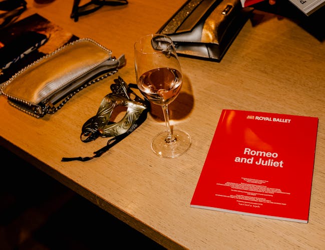 A red ballet programme for The Royal Ballet's production of Romeo and Juliet sits on a wooden table next to a wine glass of pink liquid, an ornate gold ribboned eye mask and two gold clutch bags.