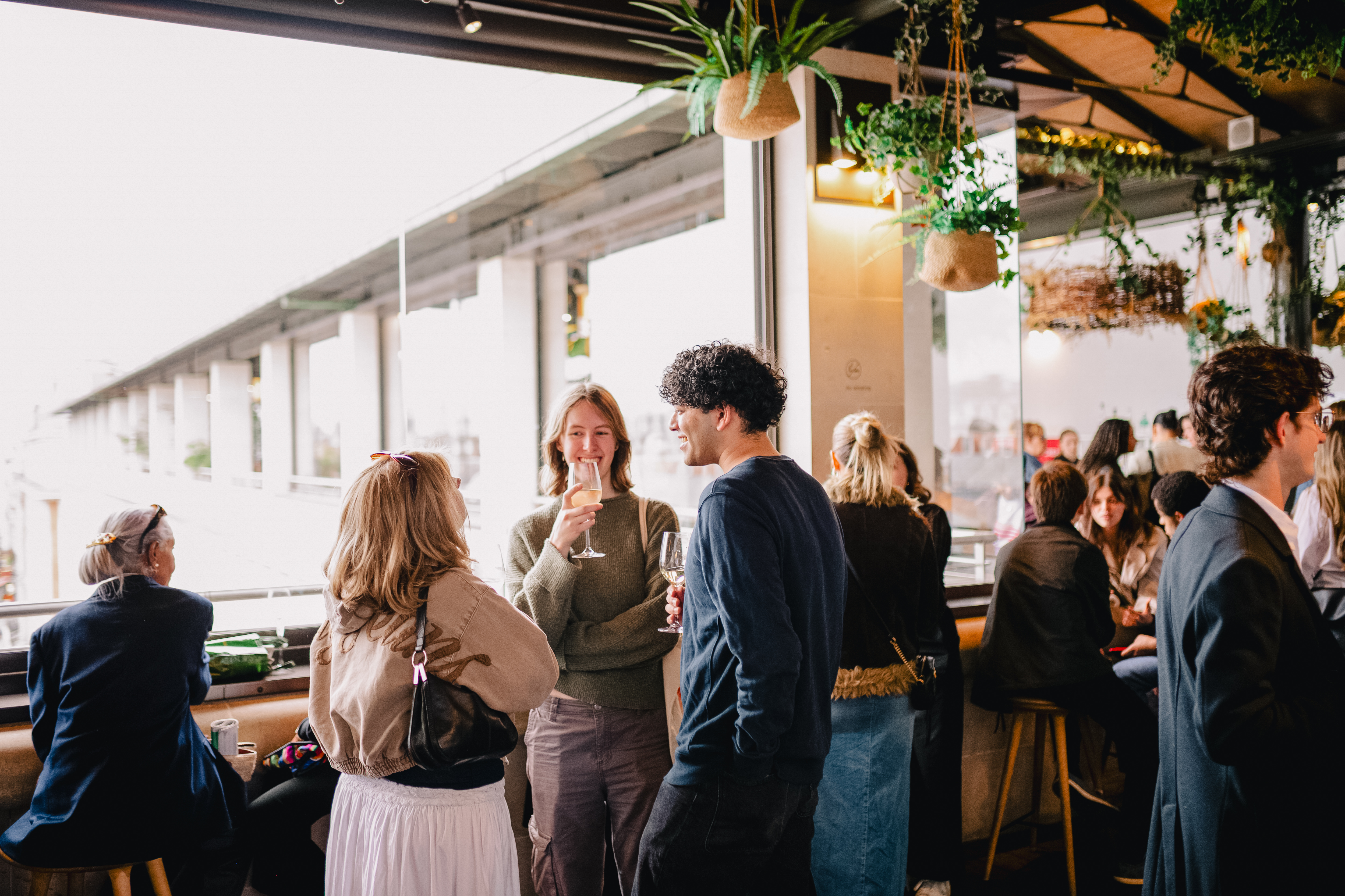A group of people stand around a terrace area of a bar in The Royal Opera House in Covent Garden. They are smiling, talking to one another and holding glasses filled with drinks. 