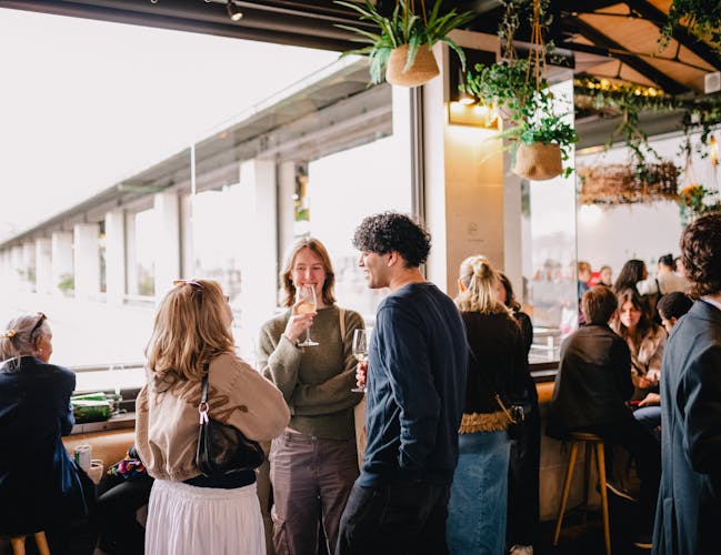 A group of people stand around a terrace area of a bar in The Royal Opera House in Covent Garden. They are smiling, talking to one another and holding glasses filled with drinks.
