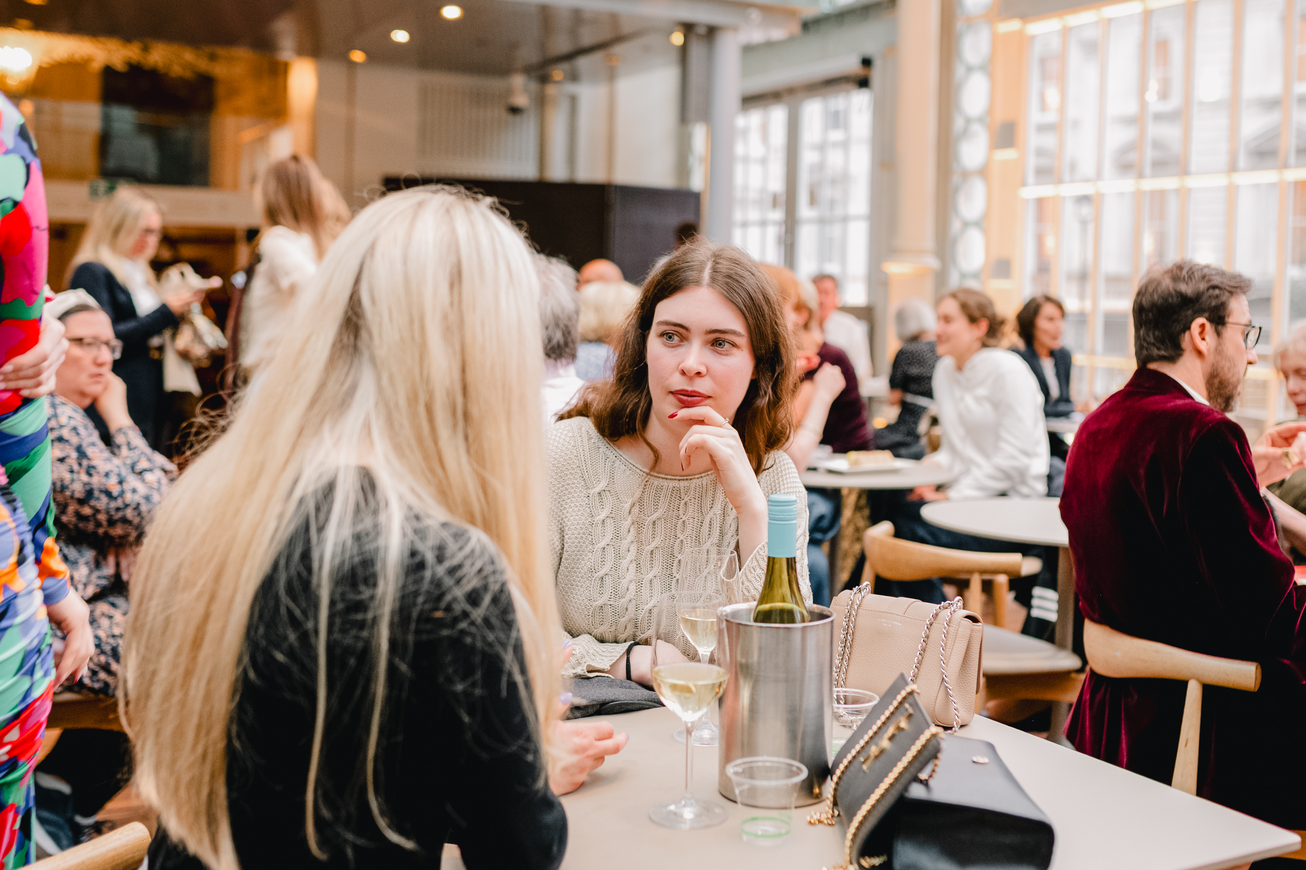 Two people are talking to one another - one is only visible from behind as their long blonde hair falls over their shoulder. The other person looks at them in conversation. They are in a busy bar area of the Paul Hamlyn Hall of the Royal Opera House in Covent Garden.