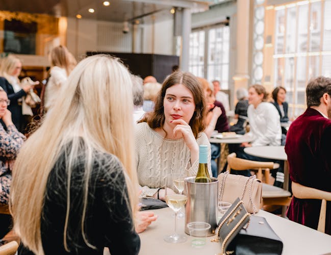Two people are talking to one another - one is only visible from behind as their long blonde hair falls over their shoulder. The other person looks at them in conversation. They are in a busy bar area of the Paul Hamlyn Hall of the Royal Opera House in Covent Garden.