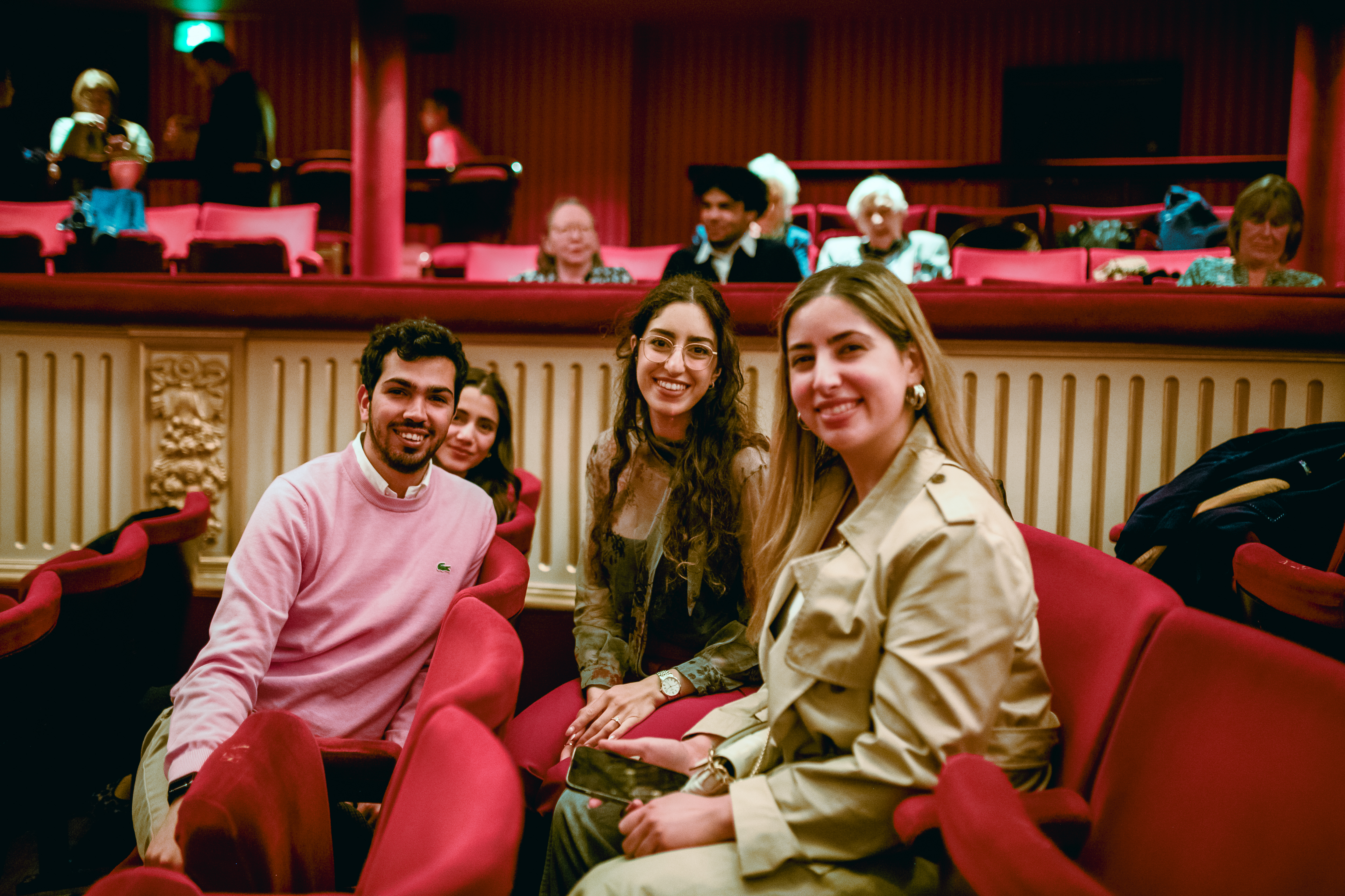 Four people sit in red velvet chairs in the auditorium of the Royal Opera House. They are smiling at the camera. 