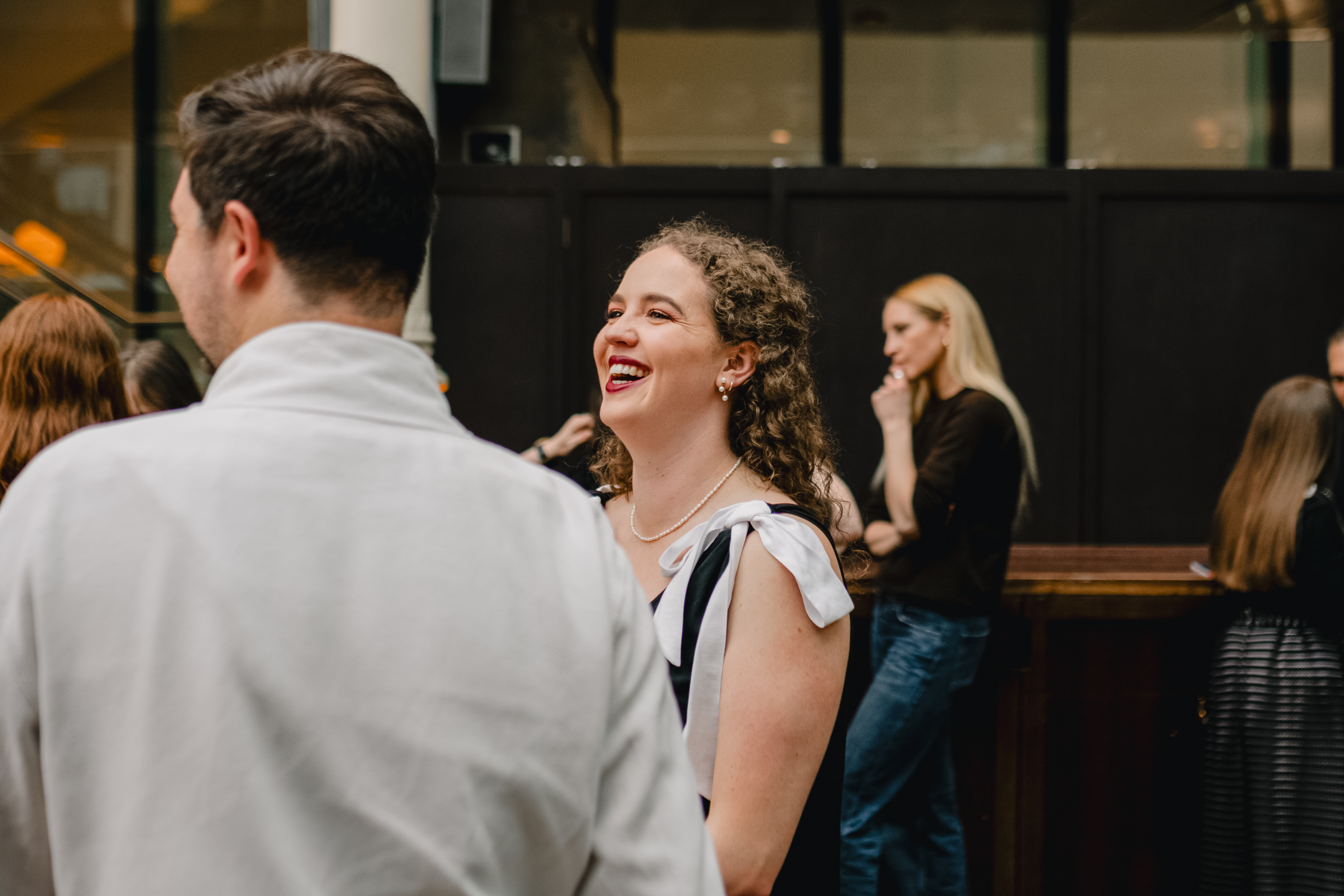 The back of someones head, their side profile slightly visible as they face left. They are turned towards another person with long, brown curly hair and they smile. Behind both of them a person with long blonde hair and long black sleeved top stands in the foreground.