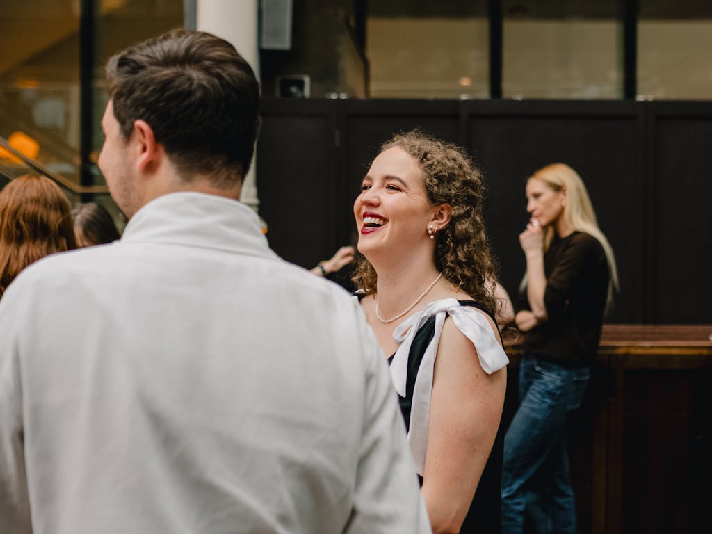 The back of someones head, their side profile slightly visible as they face left. They are turned towards another person with long, brown curly hair and they smile. Behind both of them a person with long blonde hair and long black sleeved top stands in the foreground.