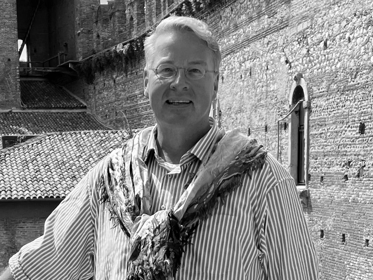 A black and white headshot of the dramaturg Alexander Meier-Dörzenbach. He stands outside leading against a railing in an old medieval style building.