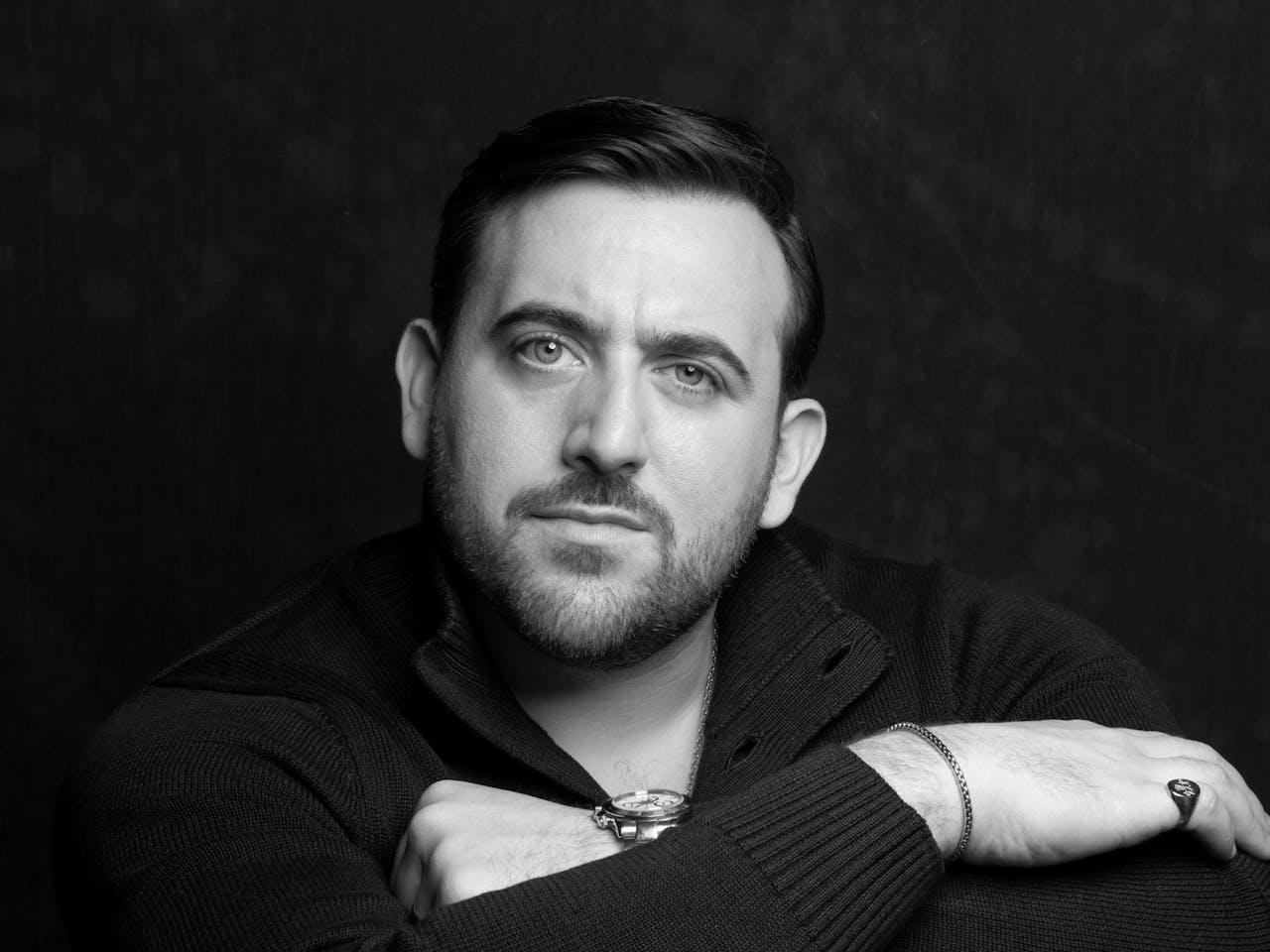 A black and white headshot of the opera singer Freddie de Tommaso. He wears a black jumper and stands against a black backdrop.