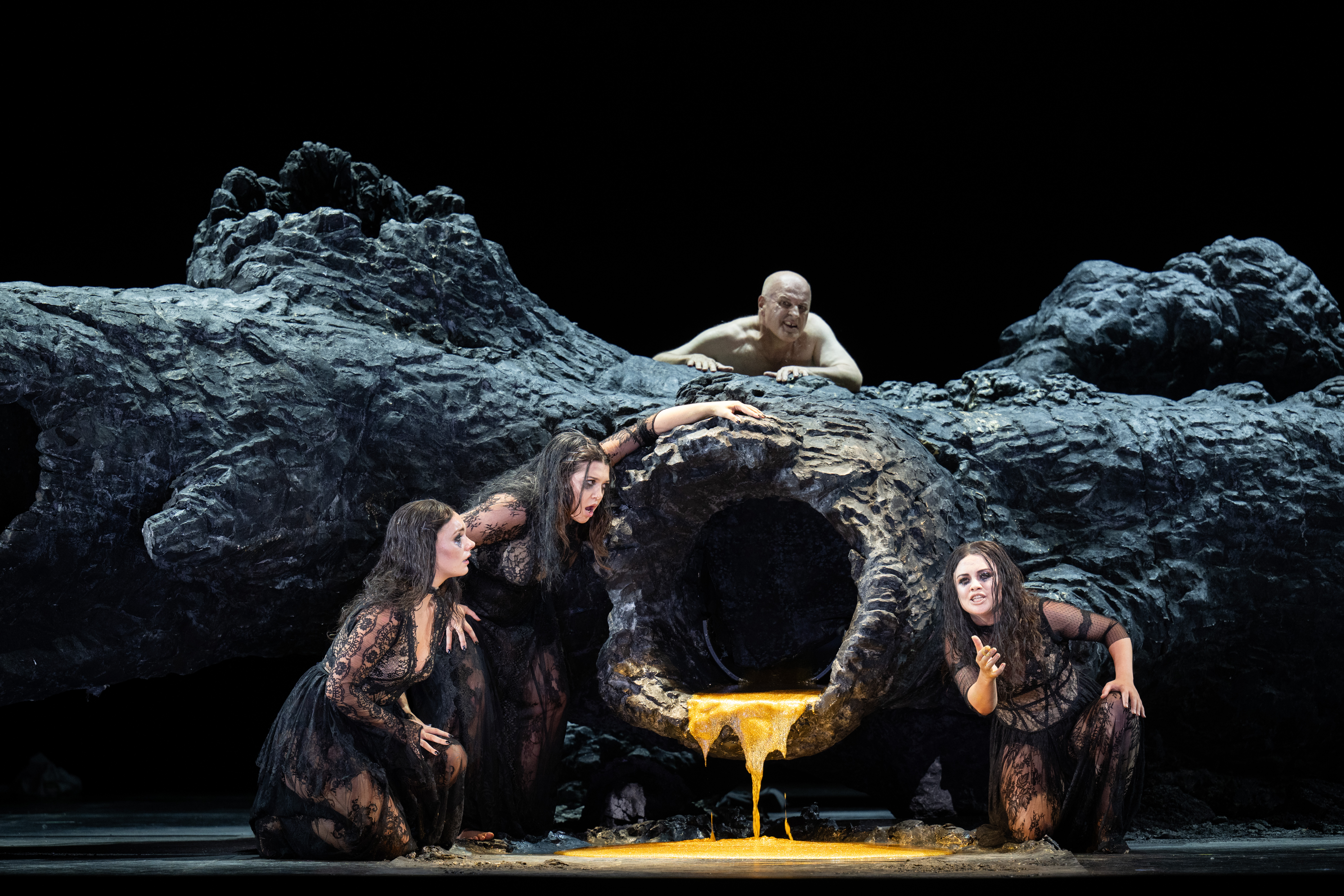 An enormous, black tree trunk is laid horizontally on a dark stage. It has a large hollow branch that faces the audience. A bright golden liquid pours from the hollow branch and spills on to the floor. Three performers in black dresses acting as Woglinde, Wellgunde and Flosshilde crouch around the flowing gold liquid. Opera singer Christopher Purves emerges from the top of the trunk and peers over, trying to catch a glimpse of the scene. (2 of 6)
