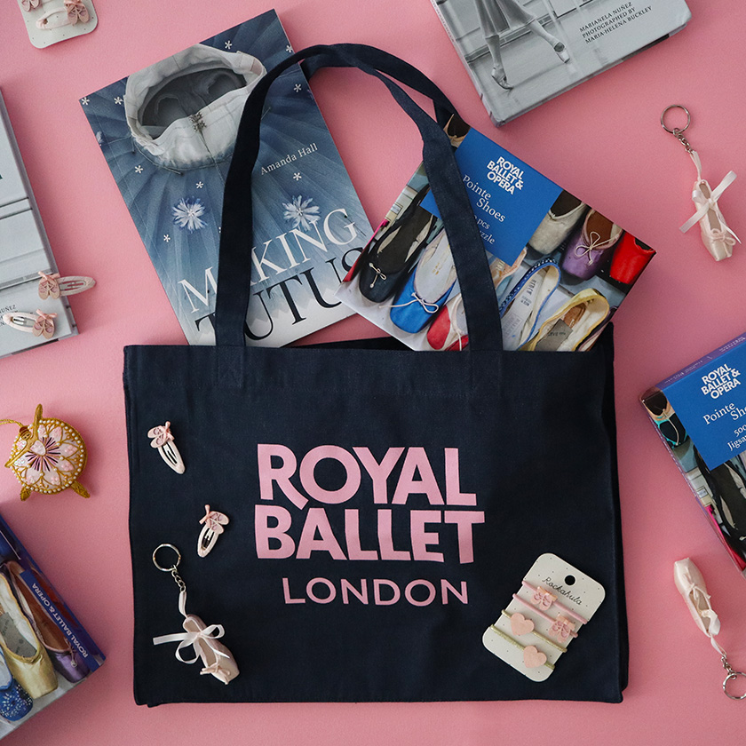 A flatlay of objects on a pink table inclduing a black tote bag that reads Royal Ballet London in pink capital letters. On front of the bag are a series of objects like keyrings. Two books emerge from the top of the bag and there are other items scattered around.